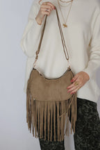 A taupe suede crescent-shaped shoulder bag with long fringe, worn crossbody by a model wearing a cream knit sweater and dark patterned pants.