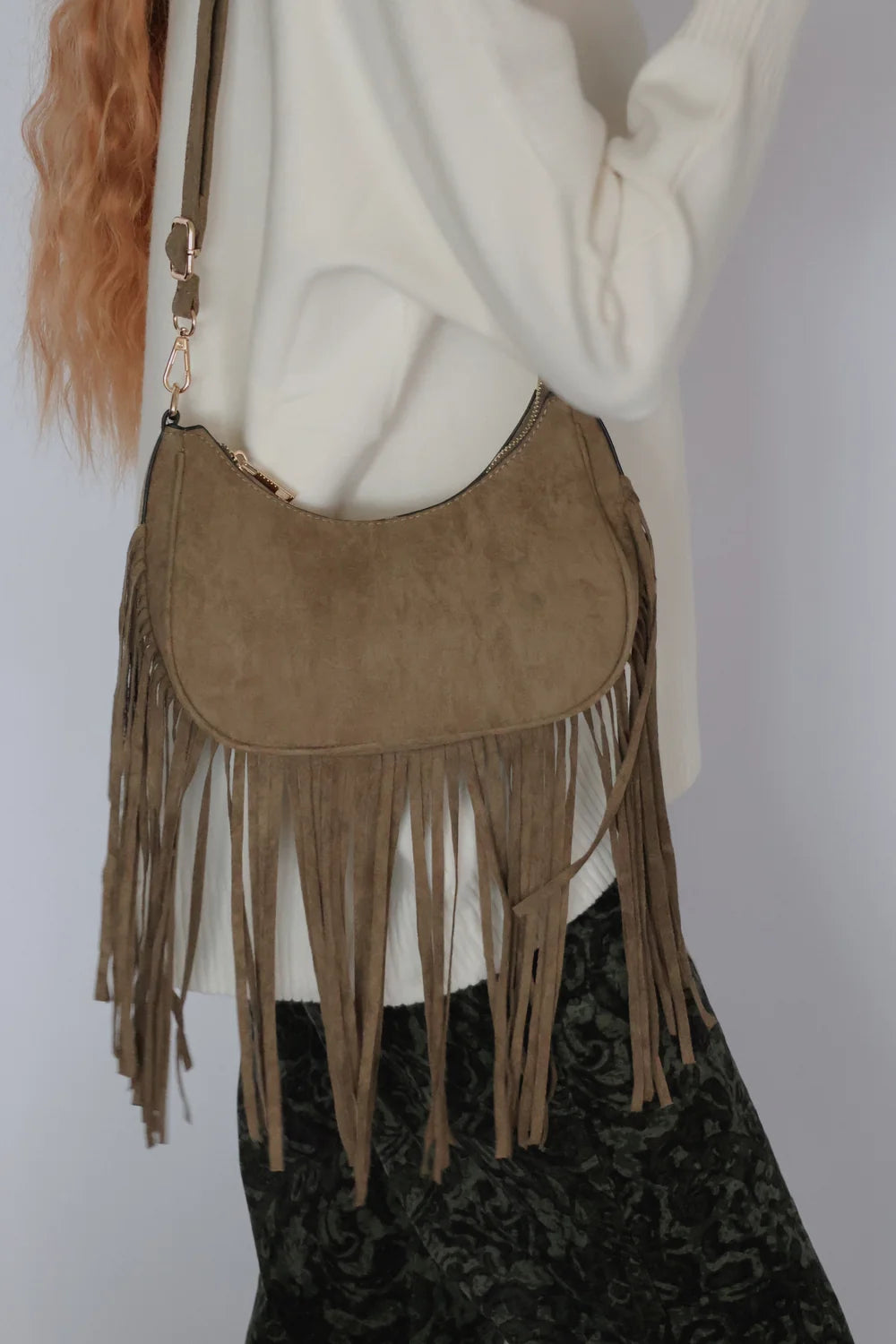 A taupe suede crescent-shaped fringe handbag worn crossbody by a person in a white sweater and dark patterned skirt, shown against a plain light gray background.