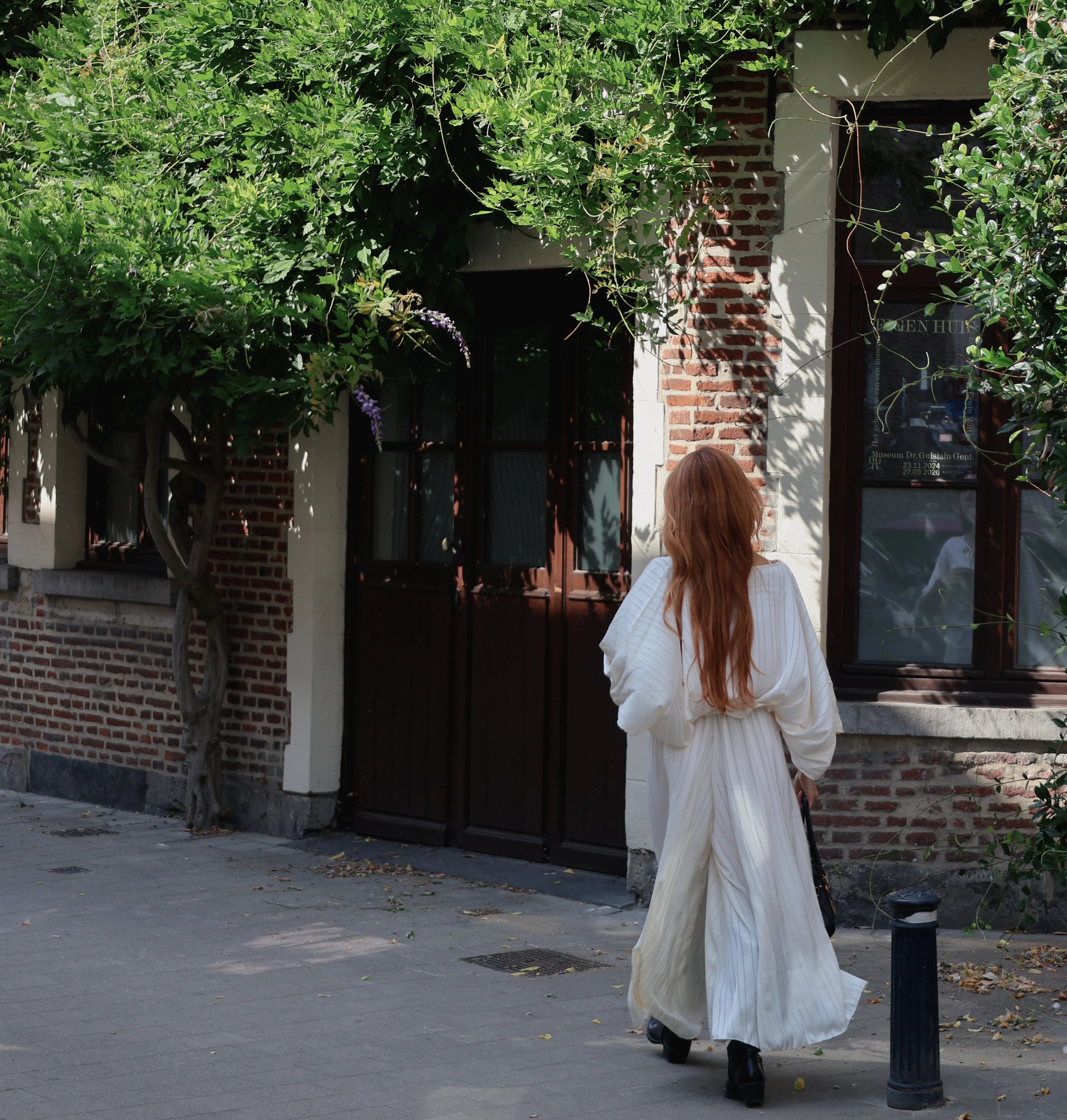 A woman with long red hair wearing a flowing white dress walks away from the camera on a sidewalk in front of a brick building with green vines covering the facade.