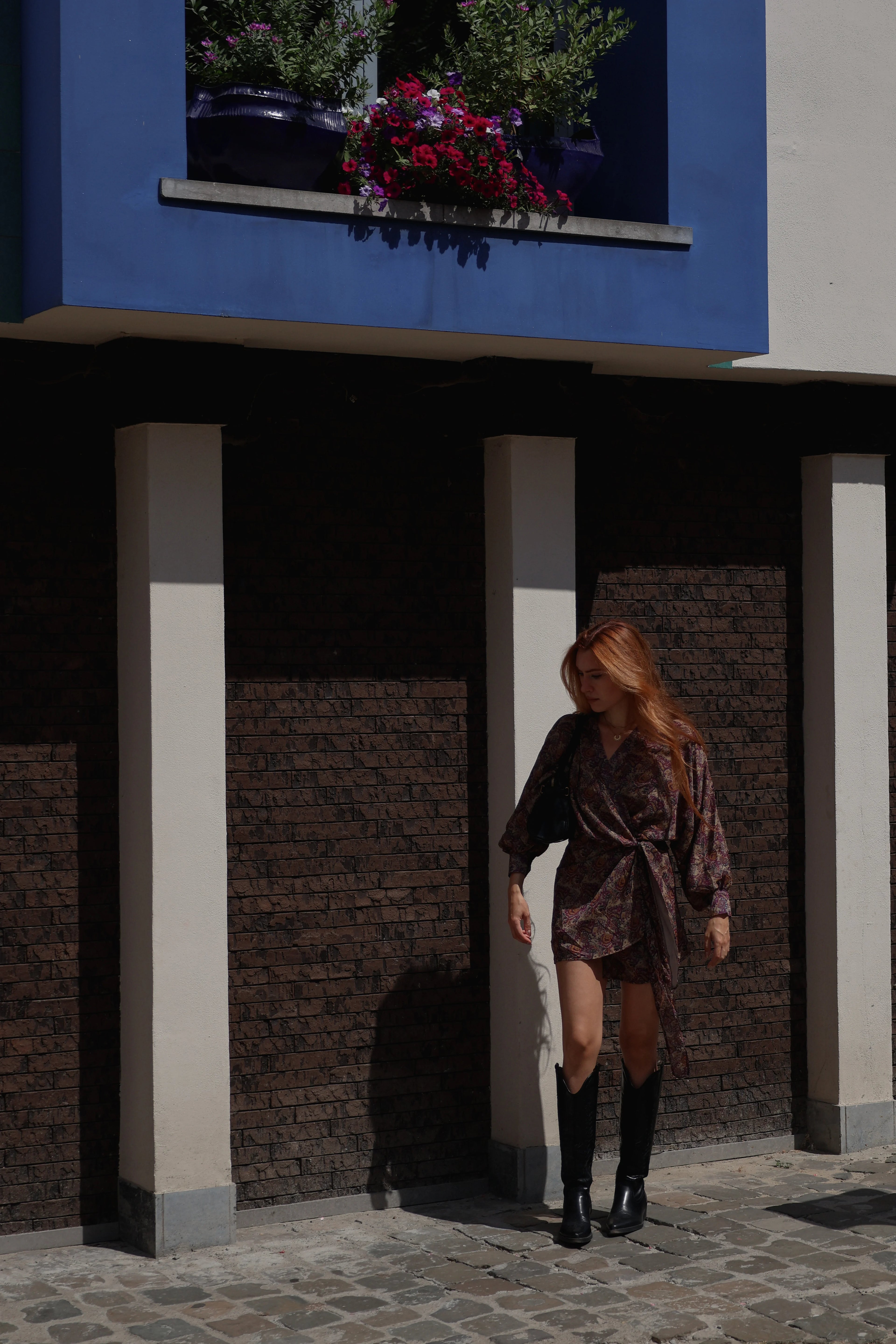 A woman with long red hair walks under a building with a blue upper facade and dark brick columns, wearing a dark brown patterned wrap dress, a black belt, and black knee-high boots.