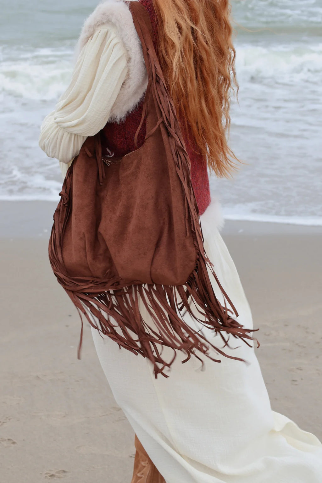 A woman on a beach wearing a brown suede fringe shoulder bag over a cream sweater and white dress, with ocean waves in the background.
