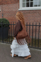 A woman with long red hair walks away from the camera on a brick sidewalk, wearing a brown fringed shoulder bag, a brown top, and a long white ruffled skirt, with a red brick building in the background.