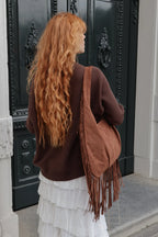A woman with long, wavy red hair is shown from behind, wearing a brown knit sweater and a white ruffled skirt while carrying a large brown suede fringe shoulder bag. She is standing in front of a dark ornate door.