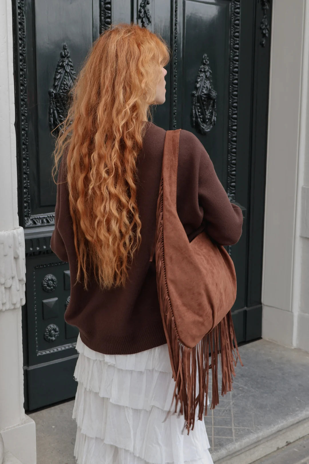 A woman with long, wavy red hair is shown from behind, wearing a brown knit sweater and a white ruffled skirt while carrying a large brown suede fringe shoulder bag. She is standing in front of a dark ornate door.