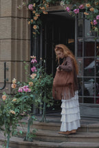 A woman with long red hair stands on the steps of a building entrance, wearing a brown fringed jacket, a long white tiered skirt, and brown boots, while holding a brown shoulder bag. The entrance is decorated with pink and orange flowers.