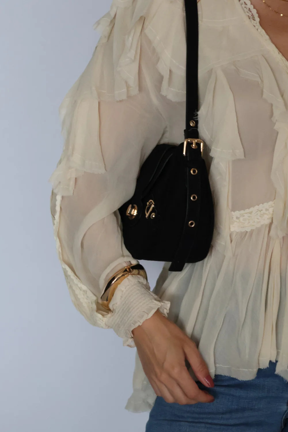 A black velvet shoulder bag with gold-tone hardware and a shoulder strap, worn by a model wearing a sheer cream ruffled blouse and blue jeans.