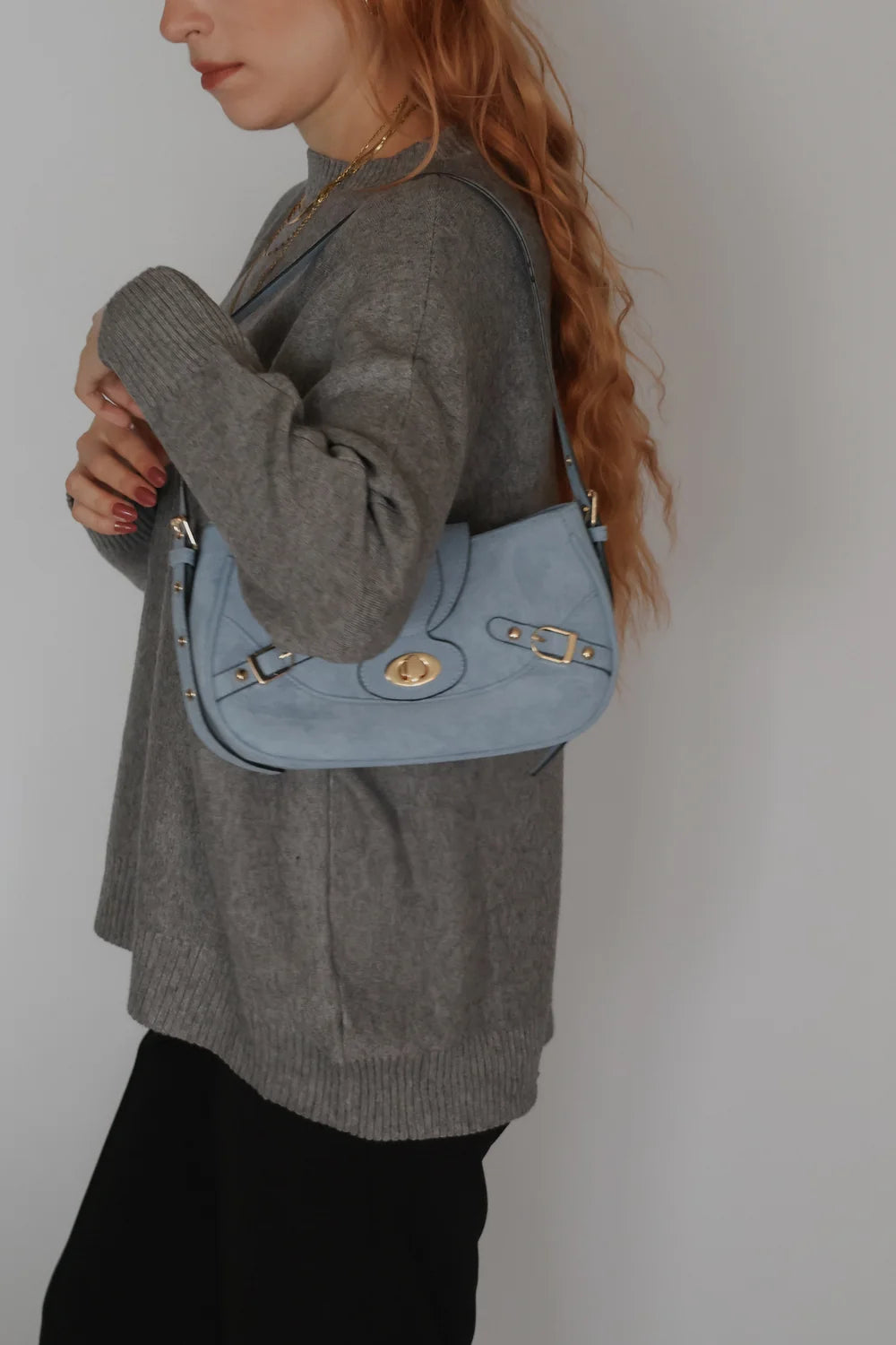 woman with red, wavy hair wears a grey sweater, a black pair of trousers, and a sky blue shoulder bag. 