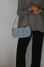 A person wearing a gray sweater and black pants carries a light blue suede shoulder bag with gold-tone hardware and a flap closure.
