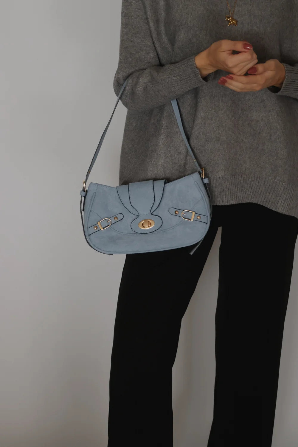 A person wearing a gray sweater and black pants carries a light blue suede shoulder bag with gold-tone hardware and a flap closure.