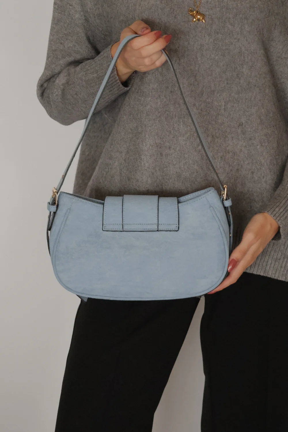 A person wearing a gray sweater and black pants holds a light blue suede crescent-shaped shoulder bag with a flap front and thin strap against a plain light gray background.