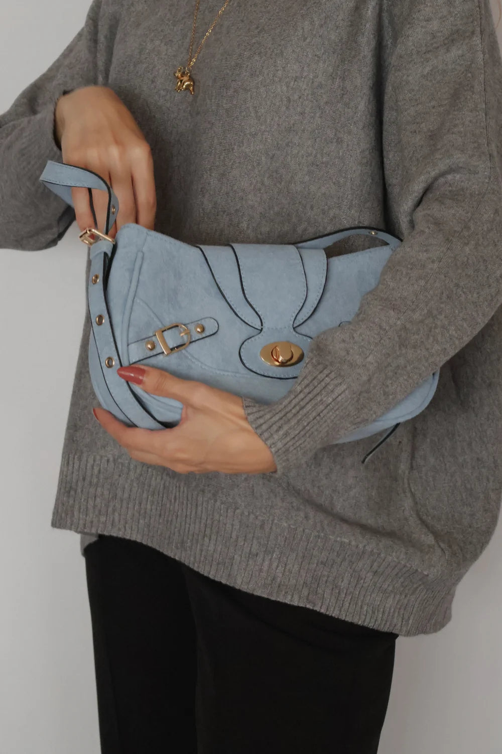 A person wearing a gray knit sweater and black pants holds a light blue suede shoulder bag with gold-tone hardware and black contrast stitching.