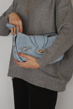 A person wearing a gray knit sweater and black pants holds a light blue suede shoulder bag with gold-tone hardware and black contrast stitching.