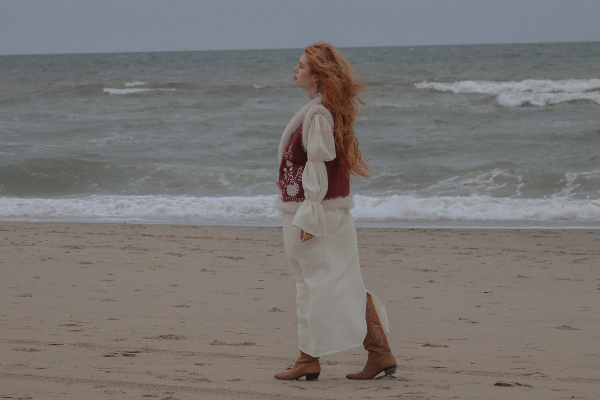 A woman with long red hair stands in profile on a sandy beach, wearing a long white dress with a dark red vest and brown cowboy boots, with the ocean waves behind her.