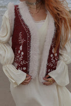 A woman on a beach wearing a cream-colored long-sleeve dress layered with a burgundy embroidered vest trimmed with white faux fur.