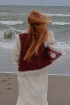 A woman with long red hair stands on a beach facing the ocean, wearing a cream-colored knit dress or robe with a burgundy vest featuring white faux-fur trim.