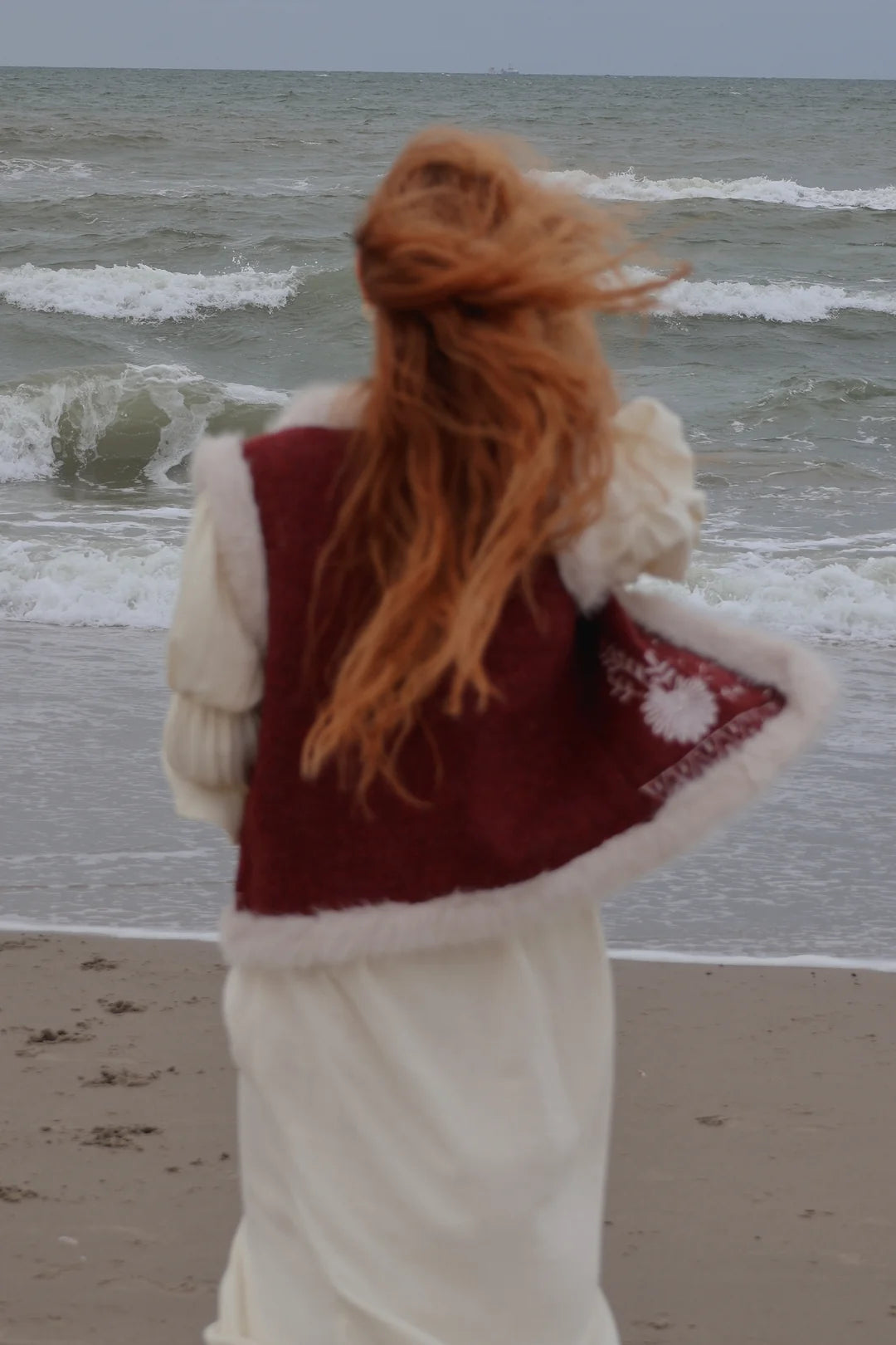 A woman with long red hair stands on a beach facing the ocean, wearing a cream-colored knit dress or robe with a burgundy vest featuring white faux-fur trim.