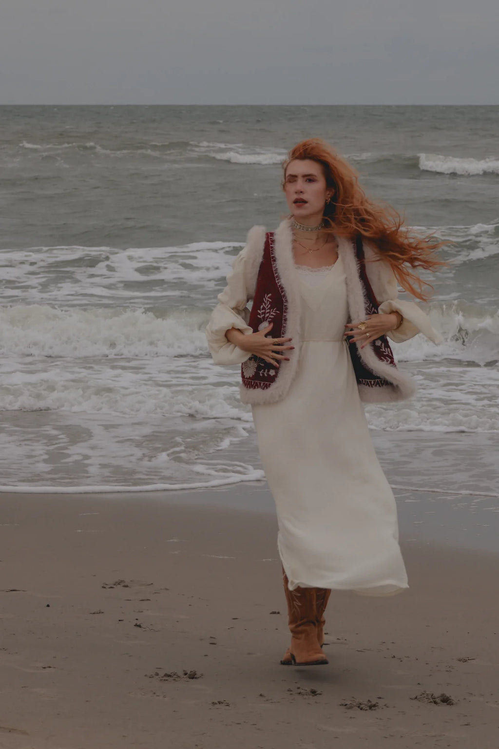 A woman with long red hair stands on a sandy beach with the ocean waves behind her, wearing a long white dress, a white faux-fur cardigan with dark red embroidered details, and brown boots.