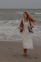 A woman with long red hair stands on a sandy beach with the ocean waves behind her, wearing a long white dress, a white faux-fur cardigan with dark red embroidered details, and brown boots.