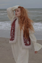 A woman with long red hair stands on a beach wearing a cream-colored dress with a burgundy embroidered vest featuring white fur trim.