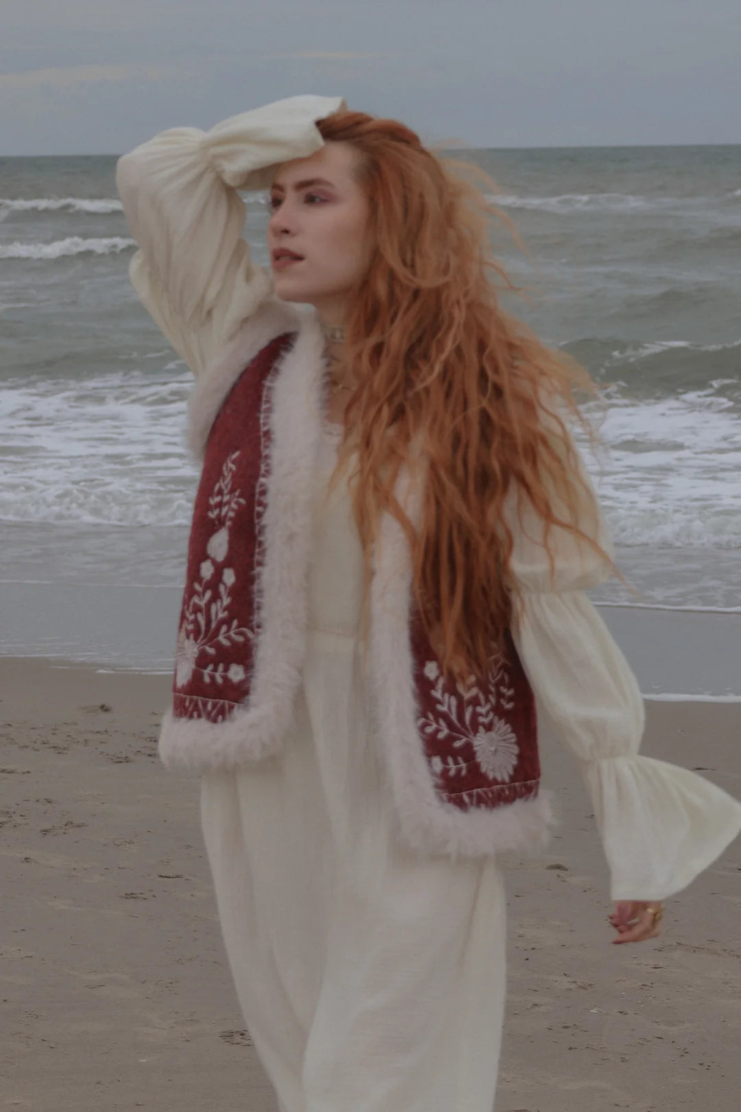 A woman with long red hair stands on a beach wearing a cream-colored dress with a burgundy embroidered vest featuring white fur trim.