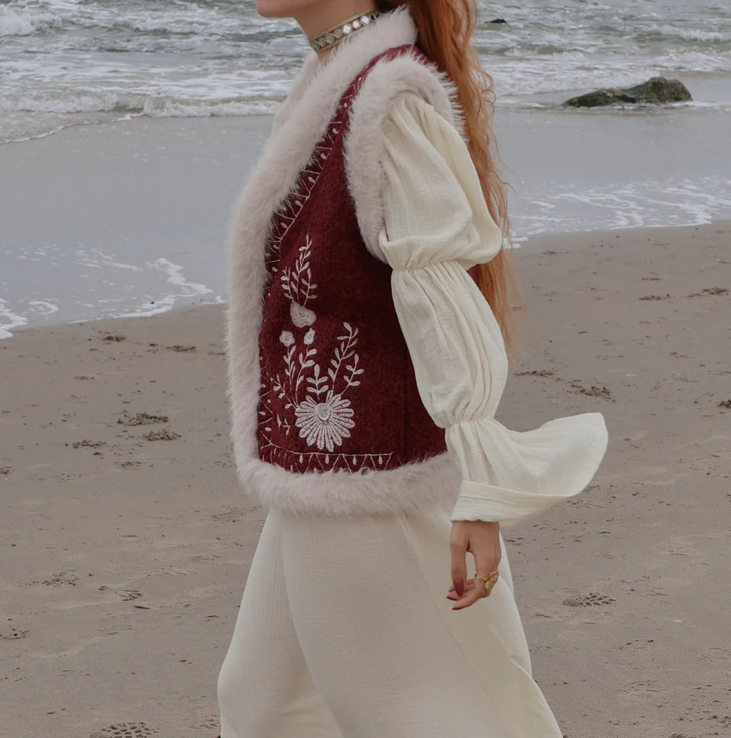 A woman wearing a burgundy embroidered vest with white faux fur trim over a cream puff-sleeve blouse and cream pants, standing on a sandy beach with the ocean in the background.