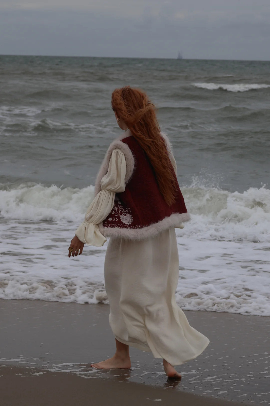 A woman with long red hair walks barefoot on a beach, wearing a cream-colored medieval-style dress with a deep burgundy vest featuring white faux-fur trim.