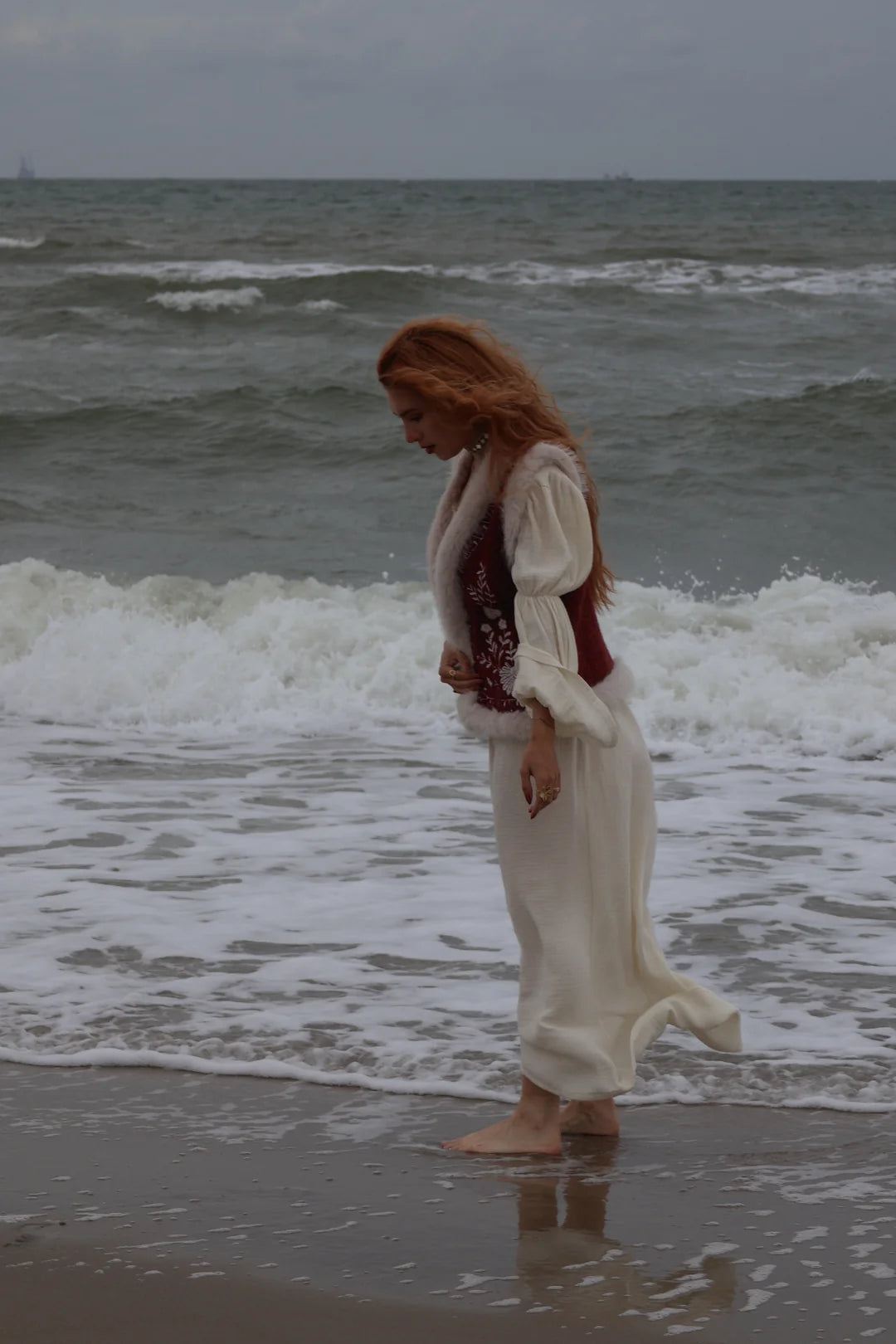 A woman with long red hair stands barefoot on a beach shoreline, wearing a flowing white dress with a dark red embroidered vest. Ocean waves are visible behind her under an overcast sky.