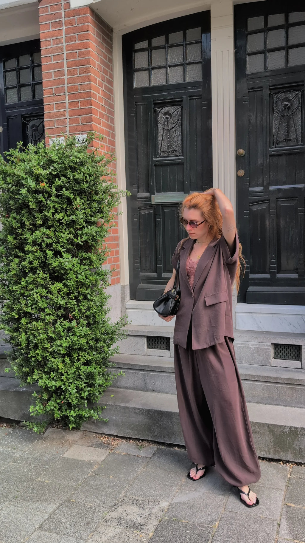 A woman with red hair wearing a matching brown two-piece suit with wide-leg pants and sunglasses, standing on a sidewalk in front of a brick building with black doors.