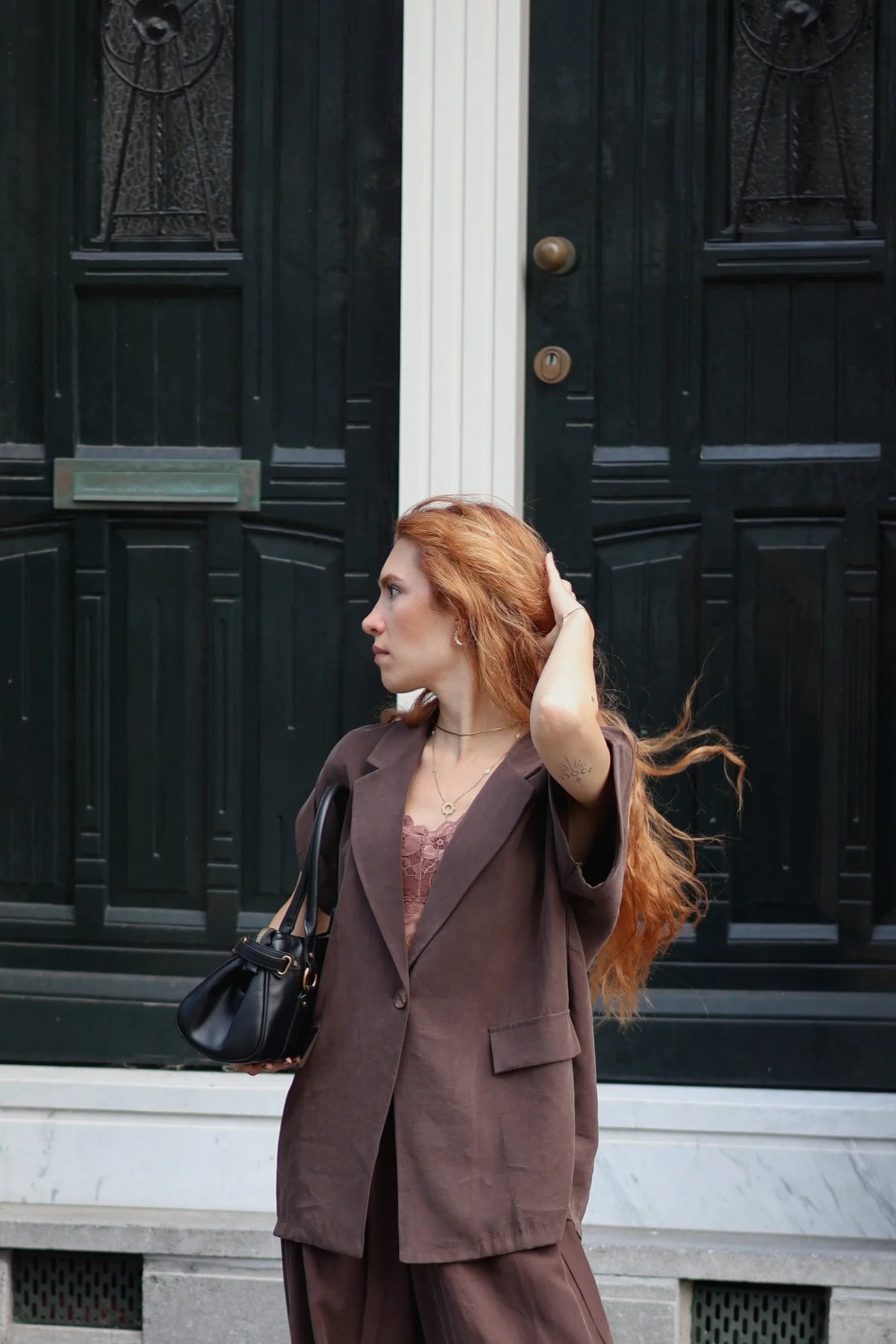 A woman with red hair stands on stone steps in front of a dark wooden door, wearing a matching brown blazer and wide-leg trousers set, a pink top, and black sandals, while holding a black handbag.