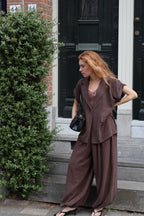 A woman with red hair stands on outdoor steps in front of a black door and brick wall, wearing a loose brown two-piece outfit with a draped top and wide-leg pants, holding a black handbag.