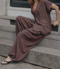 A person seated on concrete steps wearing a loose brown two-piece outfit with a short-sleeve button-front jacket and wide-leg pants, paired with black heeled sandals.