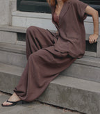 A person seated on concrete steps wearing a loose brown two-piece outfit with a short-sleeve button-front jacket and wide-leg pants, paired with black heeled sandals.