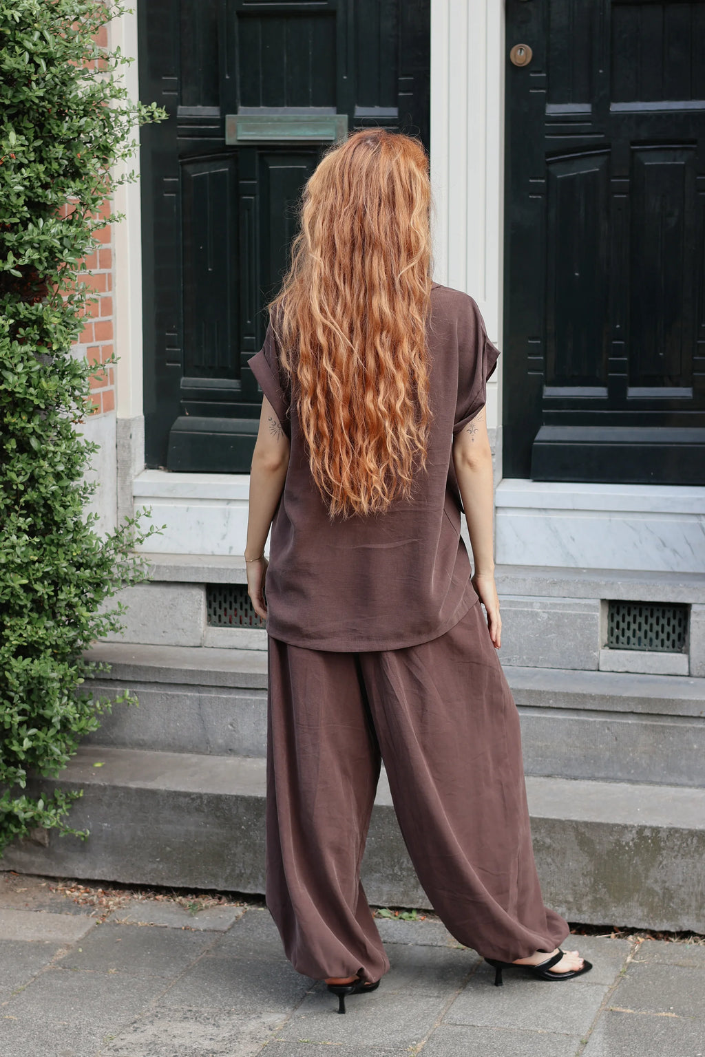 A woman with long, wavy strawberry-blonde hair is shown from behind, wearing a matching brown linen-blend tunic top and wide-leg trousers set. She is standing on a stone sidewalk in front of a building with black doors.