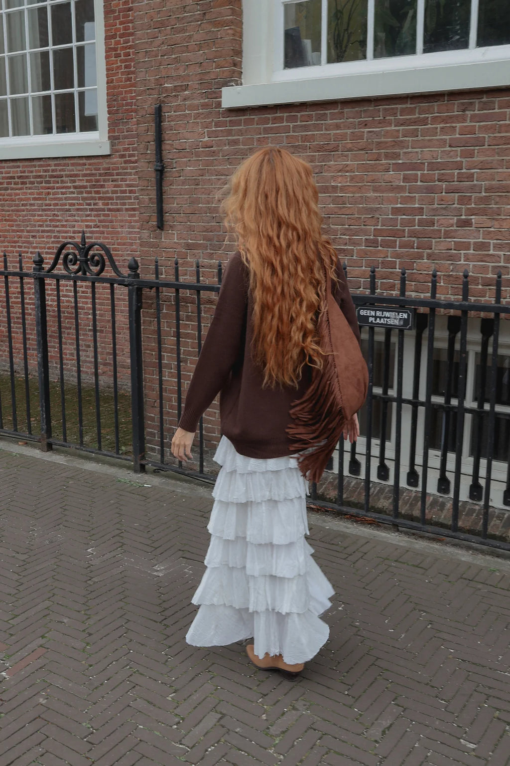 A woman with long, wavy red hair is seen from behind, wearing a brown cardigan, a white tiered ruffled maxi skirt, and carrying a brown fringed shoulder bag, standing on a brick sidewalk in front of a brick building.
