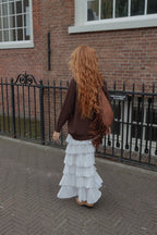 A woman with long, wavy red hair is seen from behind, wearing a brown cardigan, a white tiered ruffled maxi skirt, and carrying a brown fringed shoulder bag, standing on a brick sidewalk in front of a brick building.