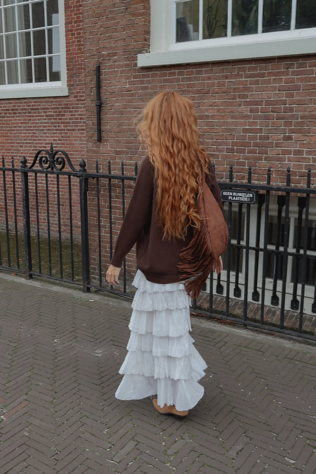 A woman with long, wavy red hair is seen from behind, wearing a brown cardigan, a white tiered ruffled maxi skirt, and carrying a brown fringed shoulder bag, standing on a brick sidewalk in front of a brick building.