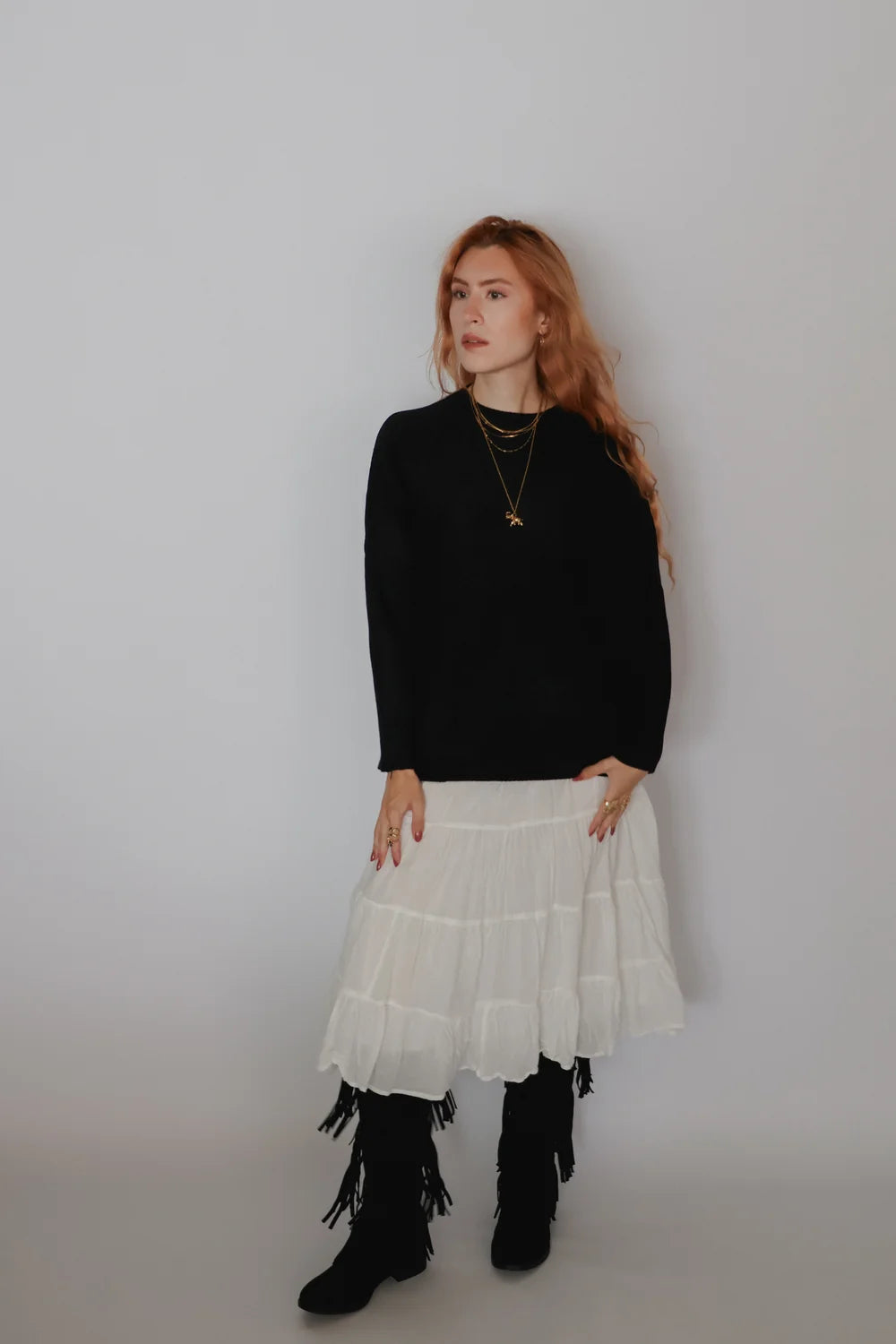 A woman stands against a plain light gray studio background wearing a black long-sleeve top, a layered white tiered skirt, black fringe boots, and layered gold necklaces.