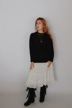 A woman stands against a plain light gray studio background wearing a black long-sleeve top, a layered white tiered skirt, black fringe boots, and layered gold necklaces.
