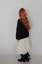 A person with long wavy red hair is shown from the back, wearing a black long-sleeve top, a tiered white skirt, and black fringed boots against a plain light gray studio background.