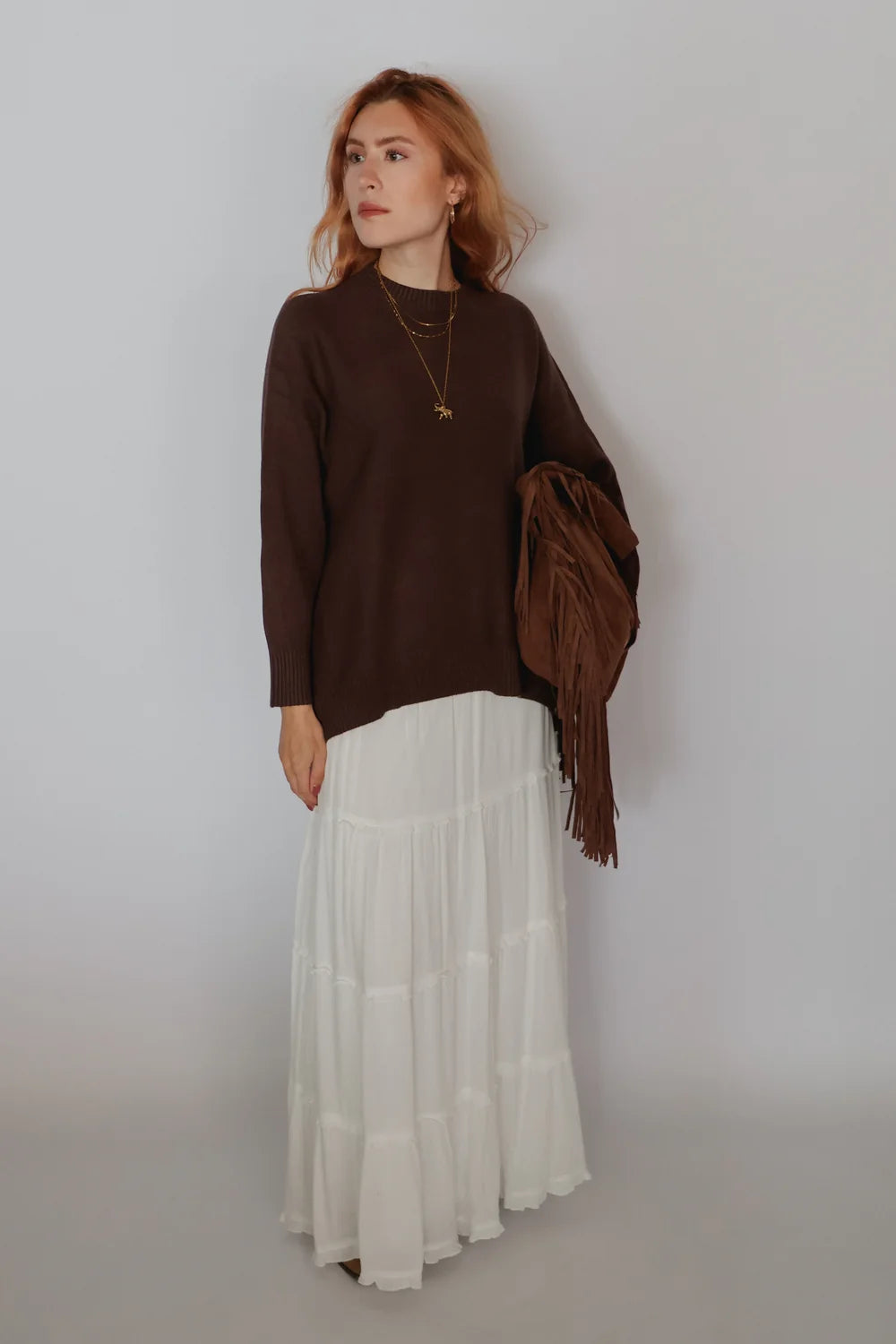 A woman stands against a plain light gray wall wearing a dark brown long-sleeve knit top and a long white tiered skirt, holding a brown fringed wrap or shawl.