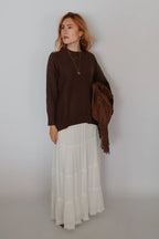 A woman stands against a plain light gray wall wearing a dark brown long-sleeve knit top and a long white tiered skirt, holding a brown fringed wrap or shawl.