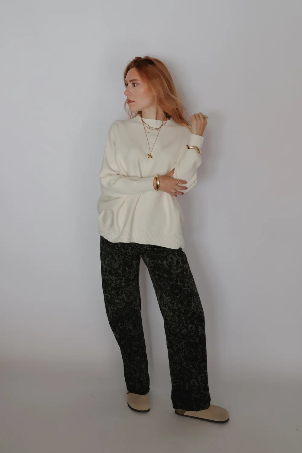 A woman stands against a plain light gray studio background wearing a cream long-sleeve top, dark patterned wide-leg pants, and beige slip-on shoes. She is posed with her arms crossed and wearing layered gold necklaces and bracelets.