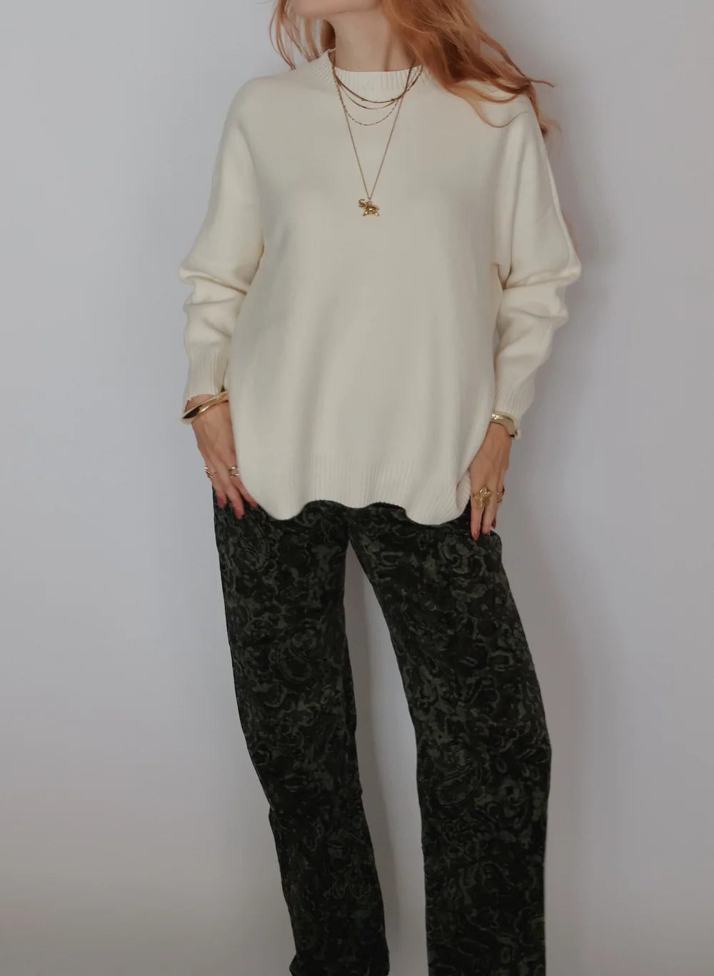 A woman wearing a cream-colored knit sweater and dark green patterned trousers, accessorized with layered gold necklaces and bracelets, standing against a plain light gray wall.