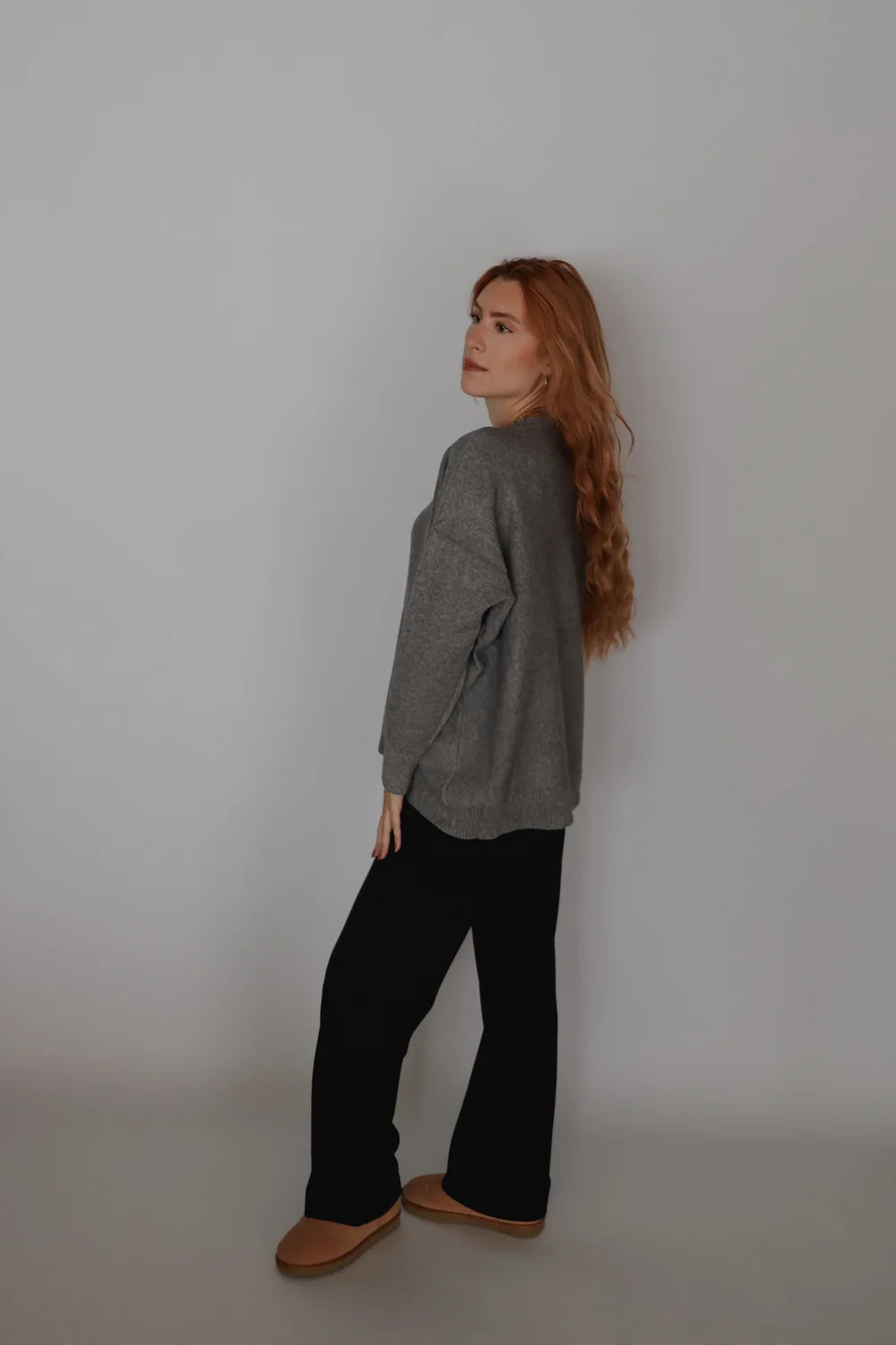 A woman with long red hair stands in profile wearing a loose gray knit sweater, black wide-leg pants, and tan slip-on shoes against a plain light gray studio background.