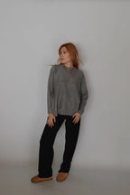 A woman with red hair stands against a plain light gray studio wall wearing a gray mock-neck knit sweater, black wide-leg pants, and tan slip-on shoes.