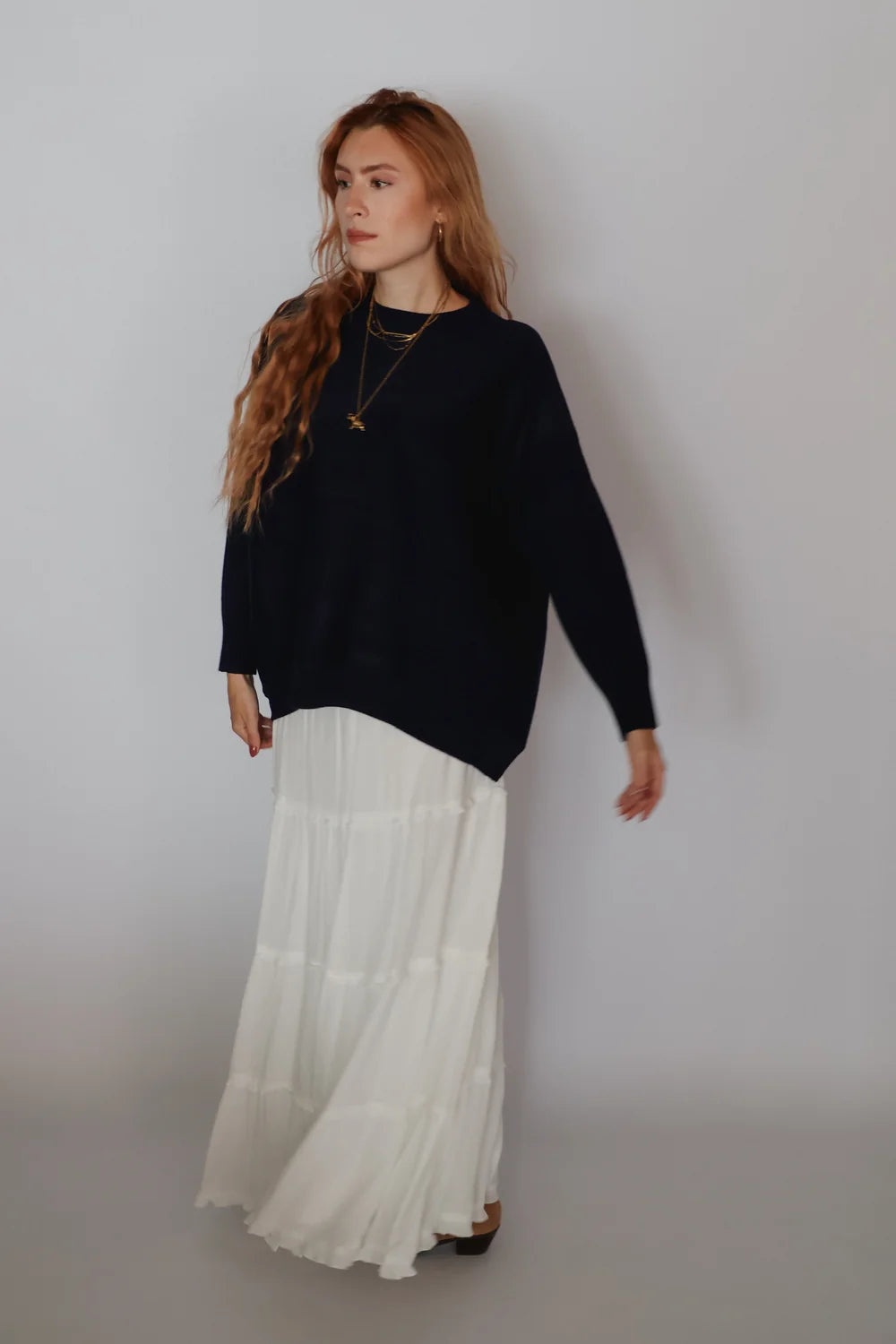A woman with long wavy light brown hair stands against a light gray studio background, wearing a loose black long-sleeve top and a long white tiered skirt. She is also wearing layered gold necklaces.