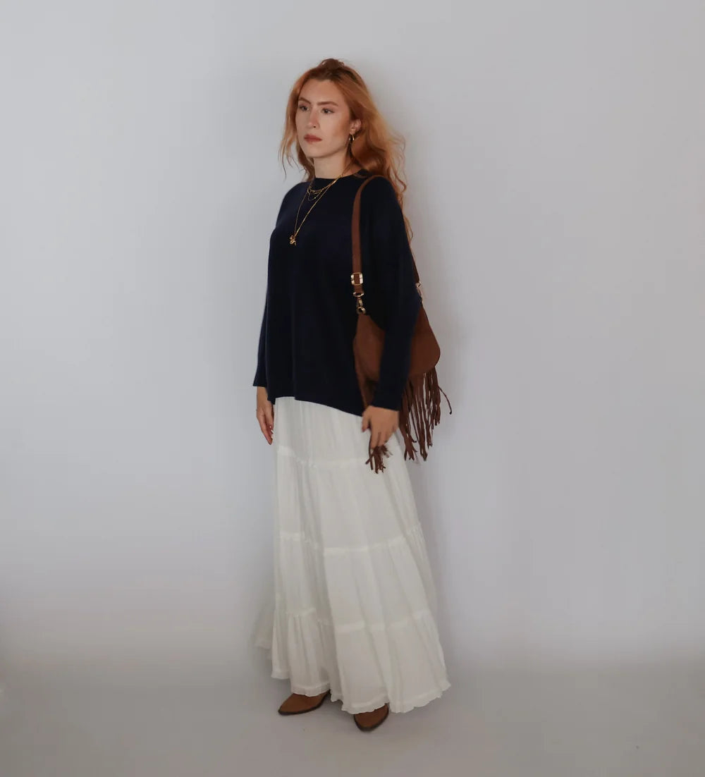 A woman stands against a plain light gray studio background wearing a dark navy long-sleeve top, a long white tiered skirt, and brown shoes, while carrying a brown fringed shoulder bag.