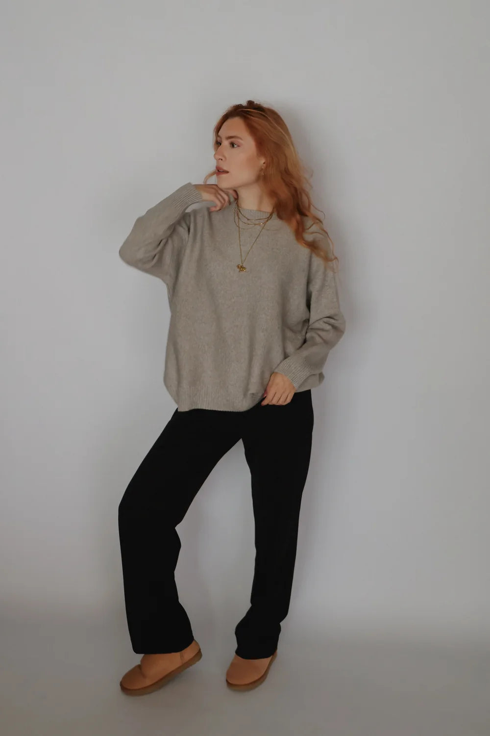 A woman with long red hair stands against a plain light gray wall wearing a loose light gray knit sweater, black pants, and tan slip-on shoes.