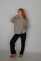 A woman with long red hair stands against a plain light gray wall wearing a loose light gray knit sweater, black pants, and tan slip-on shoes.