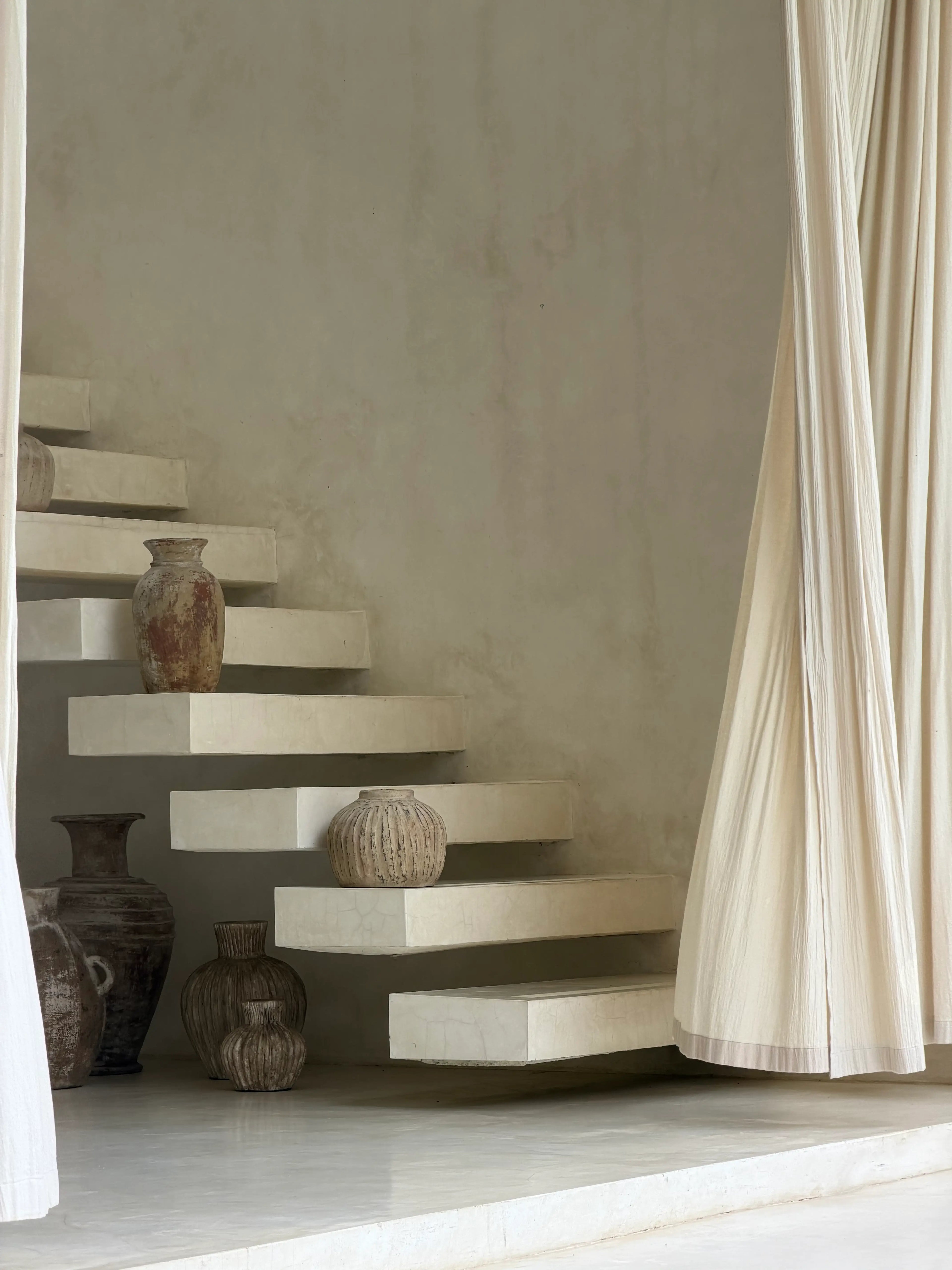 Minimalist interior scene with floating concrete stairs, draped cream fabric on both sides, and several rustic ceramic vases arranged on and beside the steps.